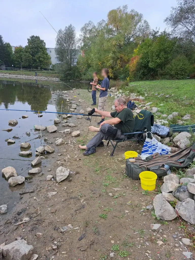 Zwei Jungangler stehen mit ihren Ruten am Ufer, vorn sitzt ein Betreuer auf dem Angelstuhl