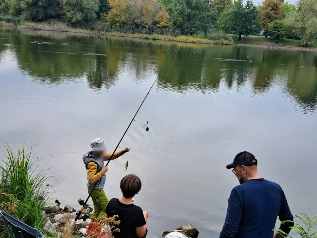 Kind mit Fischerhut hebt einen frisch gefangenen Fisch aus dem Wasser, ein Betreuer und ein weiteres Kind schauen zu