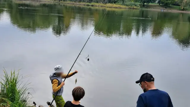Kind mit Fischerhut hebt einen frisch gefangenen Fisch aus dem Wasser, ein Betreuer und ein weiteres Kind schauen zu